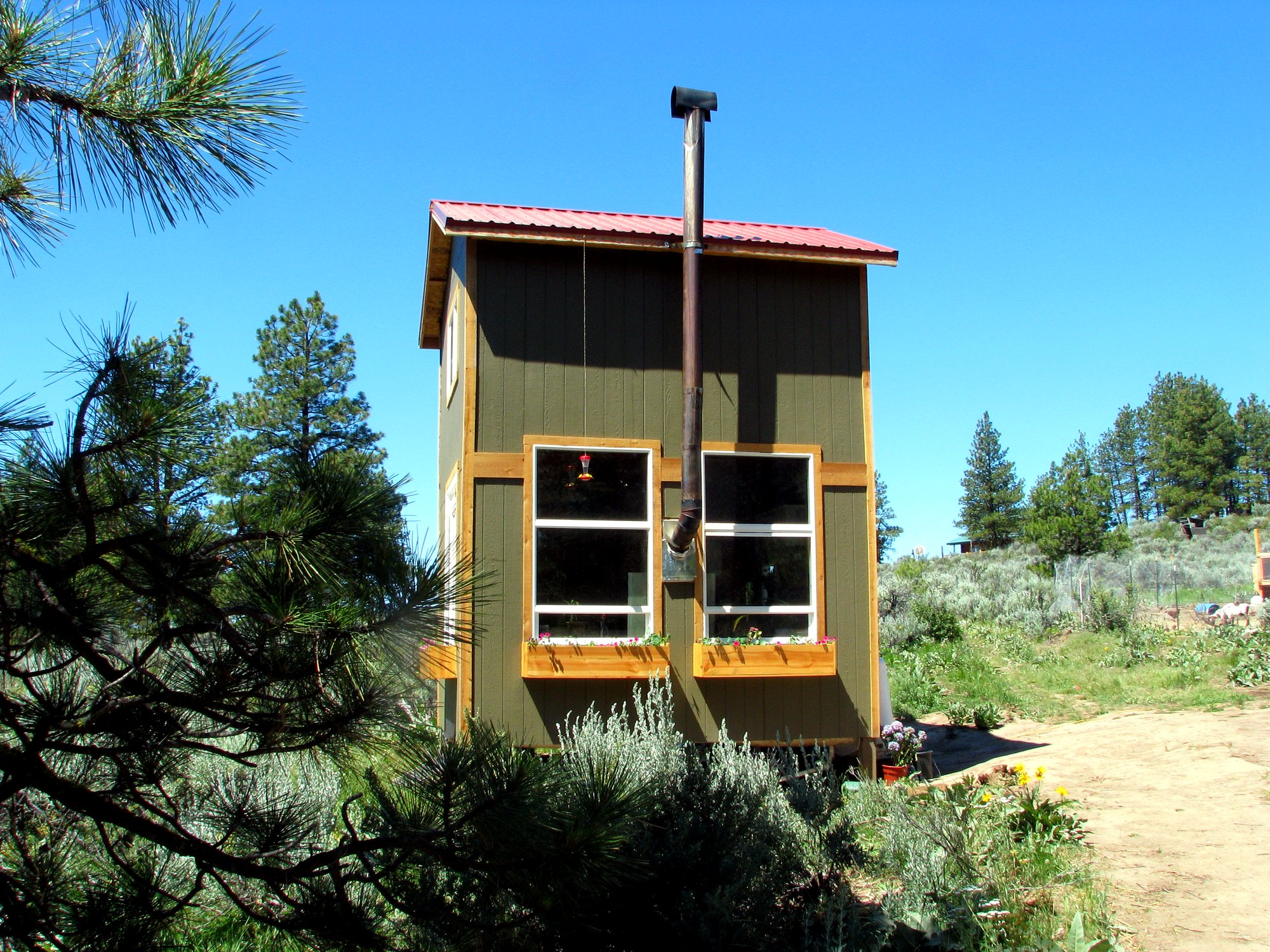 A green two-story off-grid tiny home with a red metal roof, wood-stove chimney, and flower-filled window boxes, nestled among pines