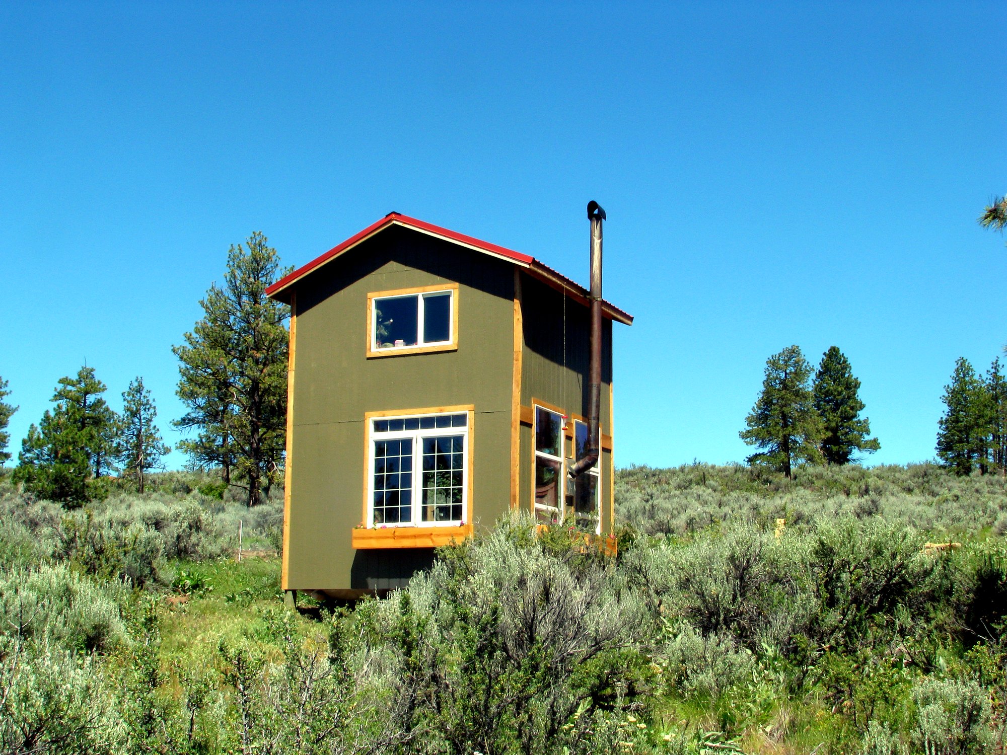 A completed two-story off-grid tiny home standing in an open high-desert meadow under a clear blue sky