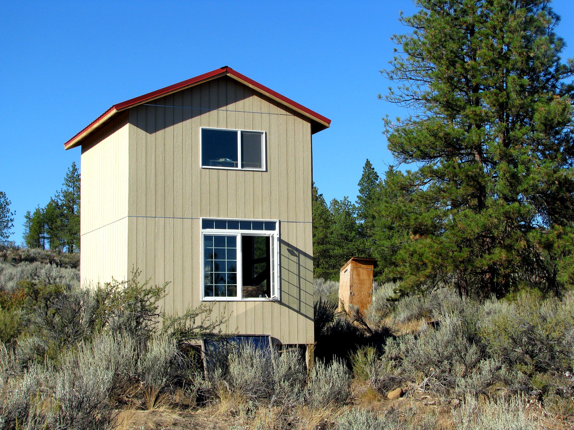 A two-story off-grid tiny home with its framed and sheathed exterior shell complete, windows installed, and red metal roof on, standing in a high-desert meadow before paint and trim